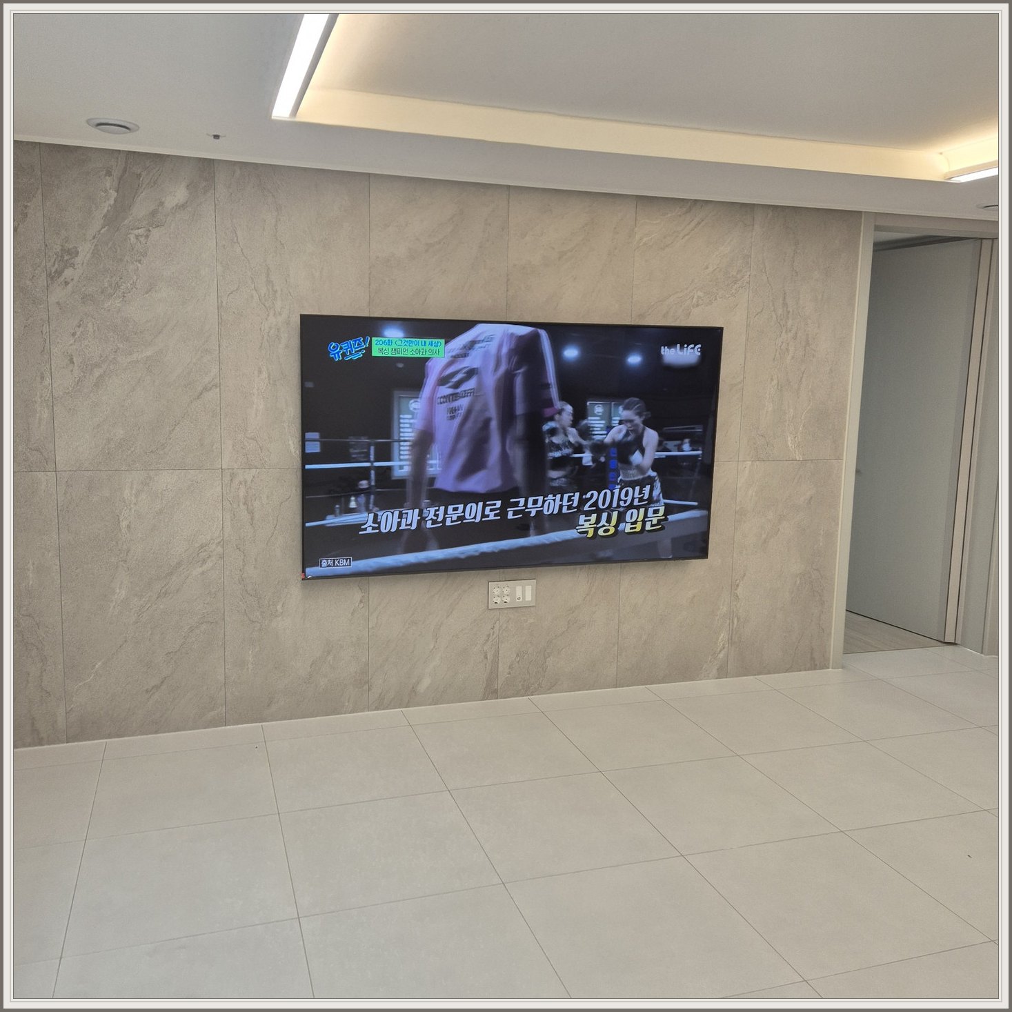 석재 느낌 벽면을 정면에서 본 TV — 벽 마감의 세로 이음선이 TV 양옆에 남아 있습니다
