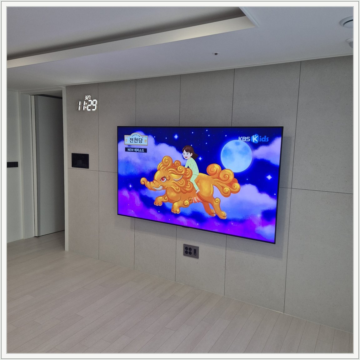 회색 판넬 벽 위의 화면과 디지털 시계 — 벽면 패널 이음선과 TV 중심의 배치를 비교할 수 있습니다