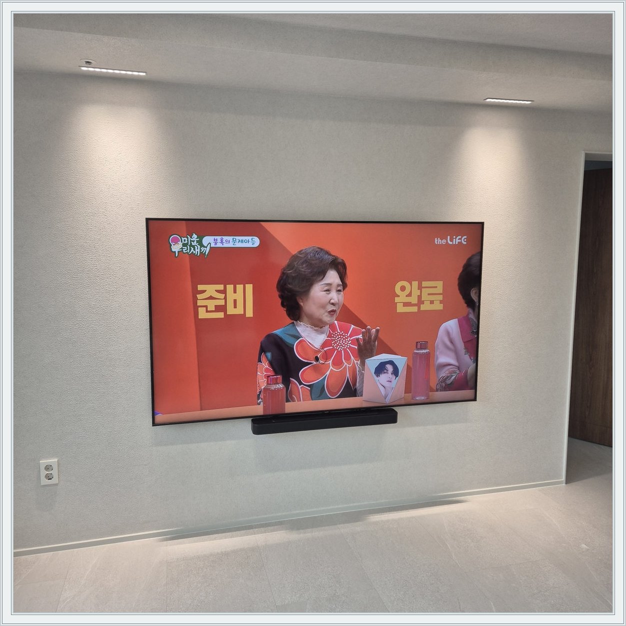 조명이 비추는 미색 벽과 사운드바 — 화면 아래 가로로 놓인 검은 음향기기가 선명합니다