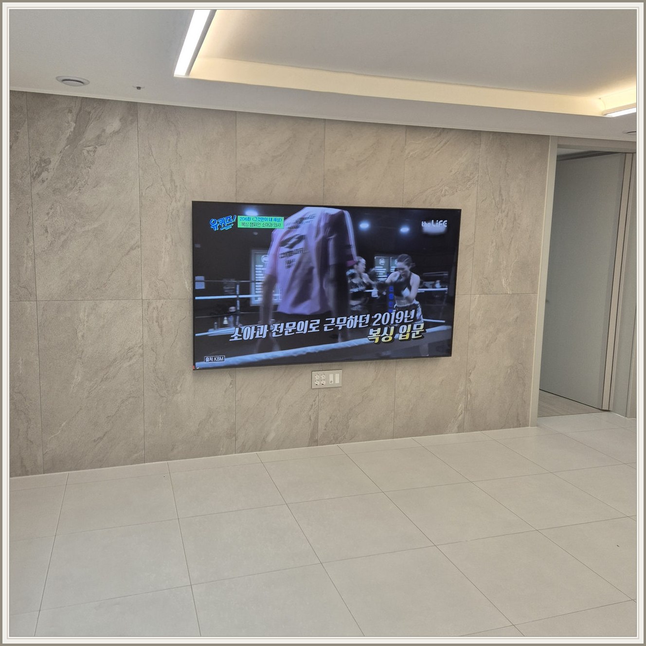 석재 느낌 벽면을 정면에서 본 TV — 넓은 바닥 여유와 조명 라인이 동시에 보입니다