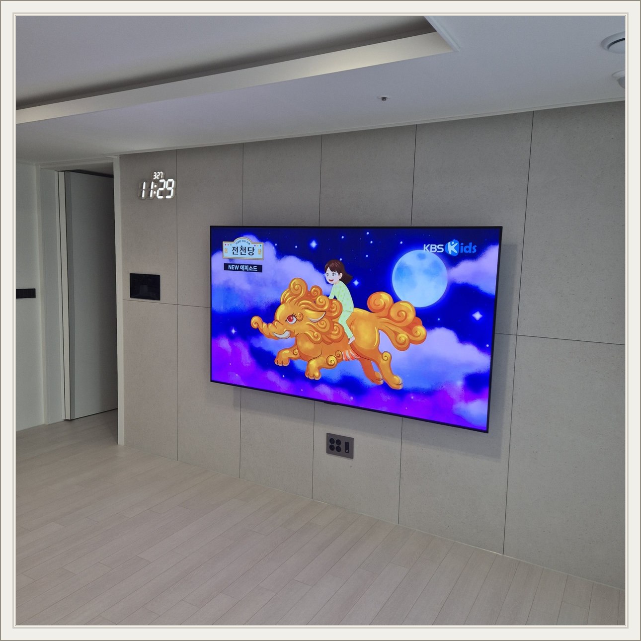 회색 판넬 벽 위의 화면과 디지털 시계 — 벽면 패널 이음선과 TV 중심의 배치를 비교할 수 있습니다