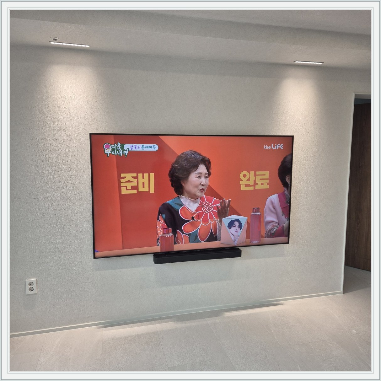 조명이 비추는 미색 벽과 사운드바 — 화면 아래 가로로 놓인 검은 음향기기가 선명합니다