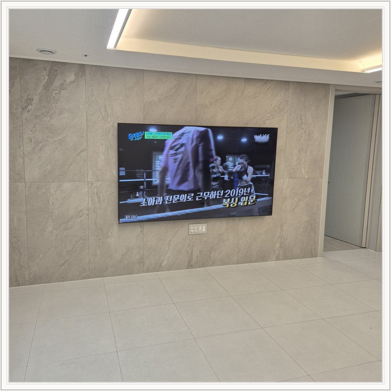 석재 느낌 벽면을 정면에서 본 TV — 넓은 바닥 여유와 조명 라인이 동시에 보입니다