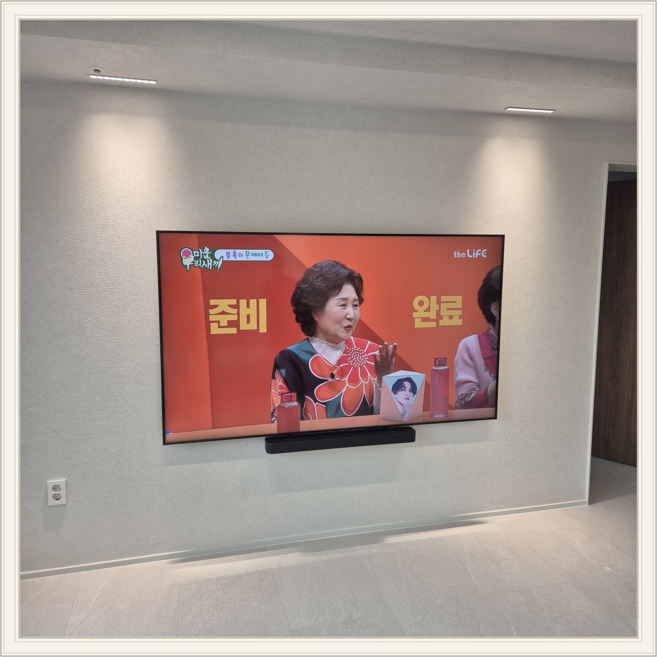 조명이 비추는 미색 벽과 사운드바 — 화면 아래 가로로 놓인 검은 음향기기가 선명합니다