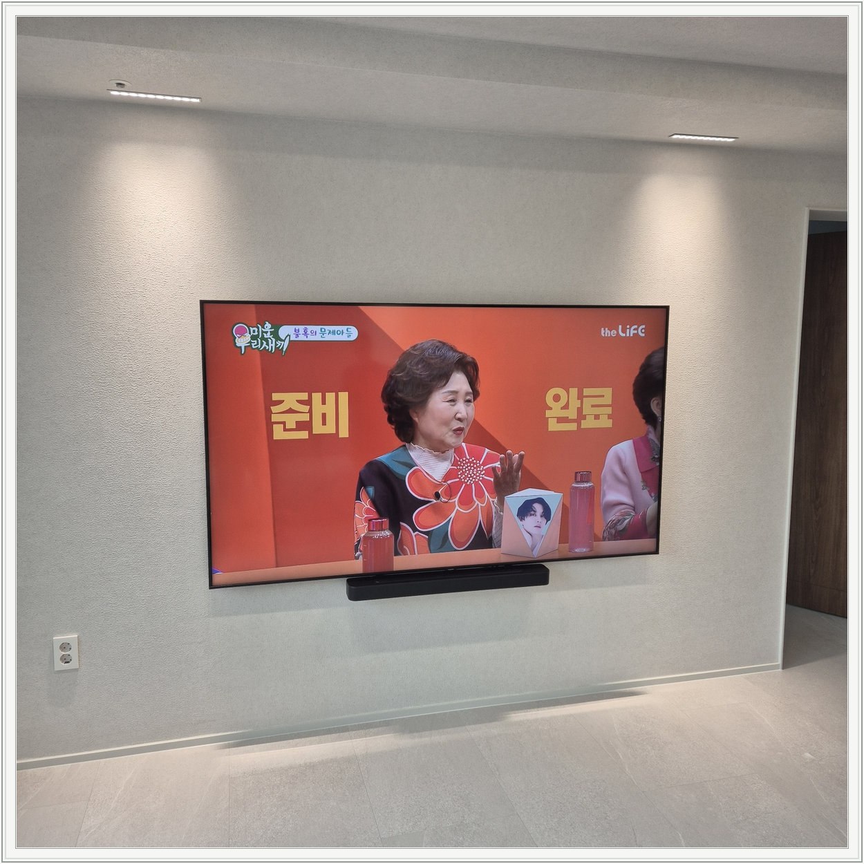 조명이 비추는 미색 벽과 사운드바 — 화면 아래 가로로 놓인 검은 음향기기가 선명합니다